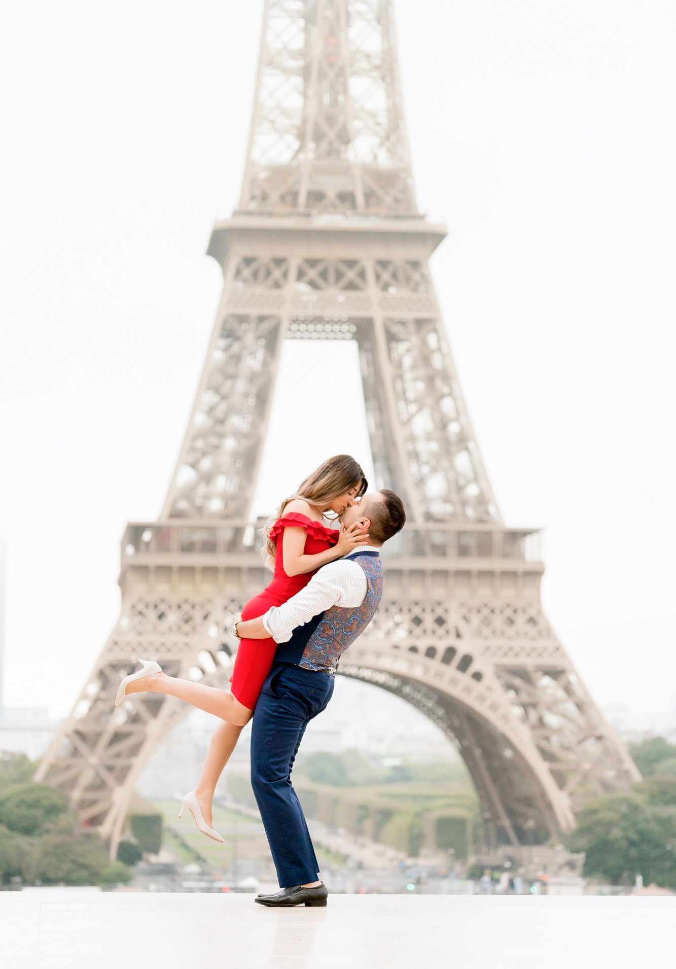 Man in a blue suit and vest lifting a woman in a red off-the-shoulder dress for a kiss in front of the Eiffel Tower during a professional Paris couple's portrait session at Trocadero.