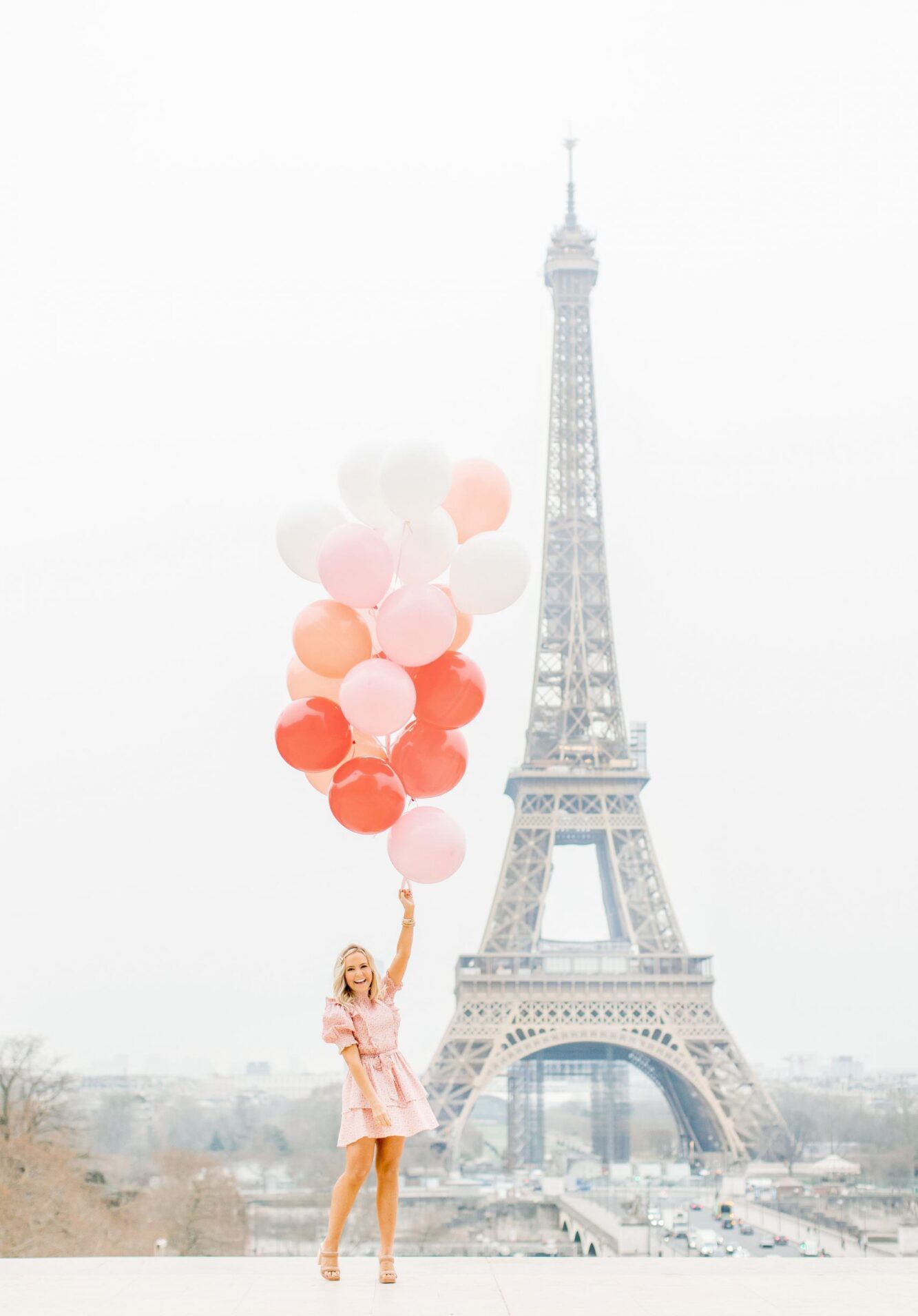 A solo woman in a pink dress holding a large bouquet of pink and white balloons at Trocadéro during an individual photoshoot with a Paris photographer. The Eiffel Tower stands tall in the background under a soft, bright sky.