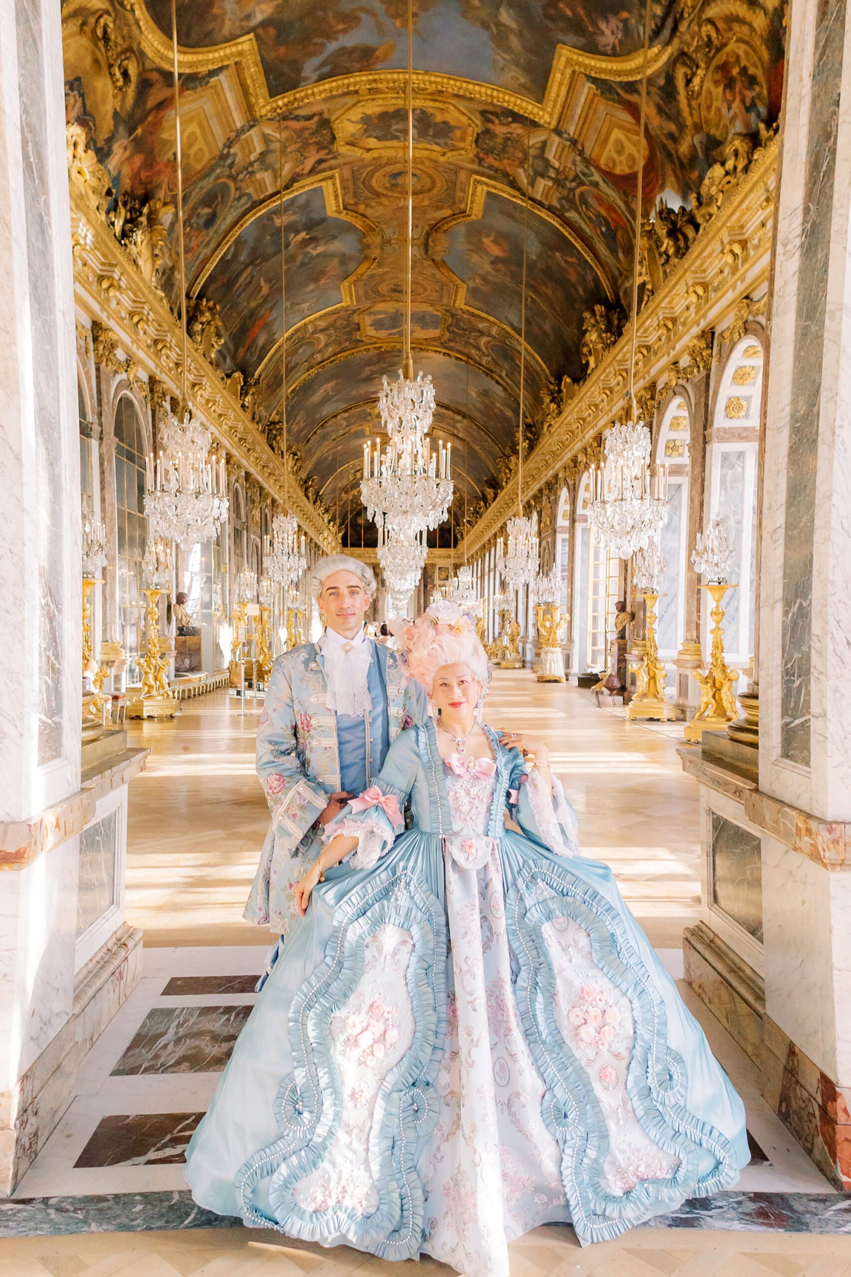 Full-length portrait of a couple in stunning historical costumes inside the Hall of Mirrors at the Palace of Versailles