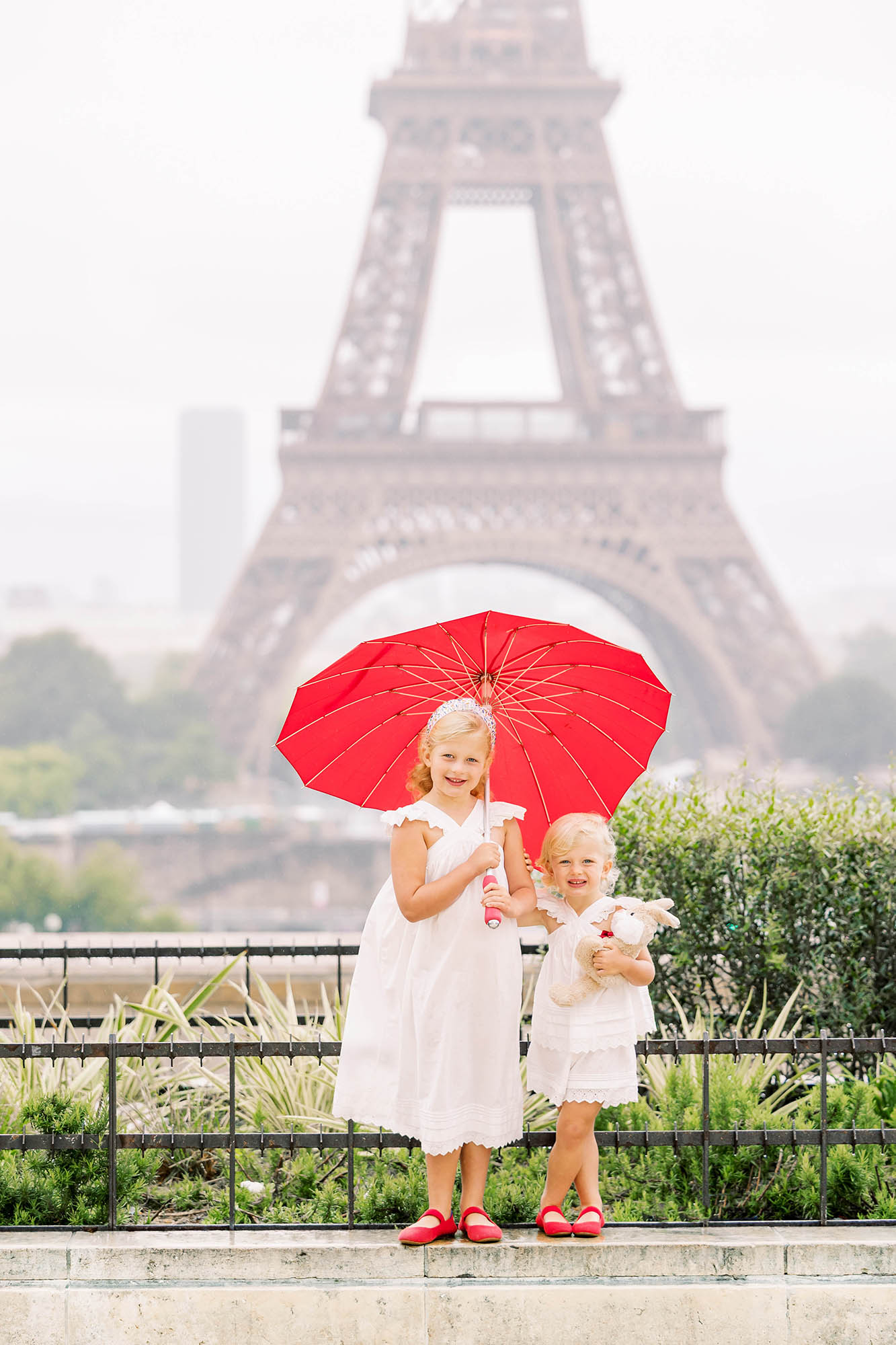 cute photo of two young sisters holding a red heart umbrella in front of the eiffel tower in paris during their family photoshoot