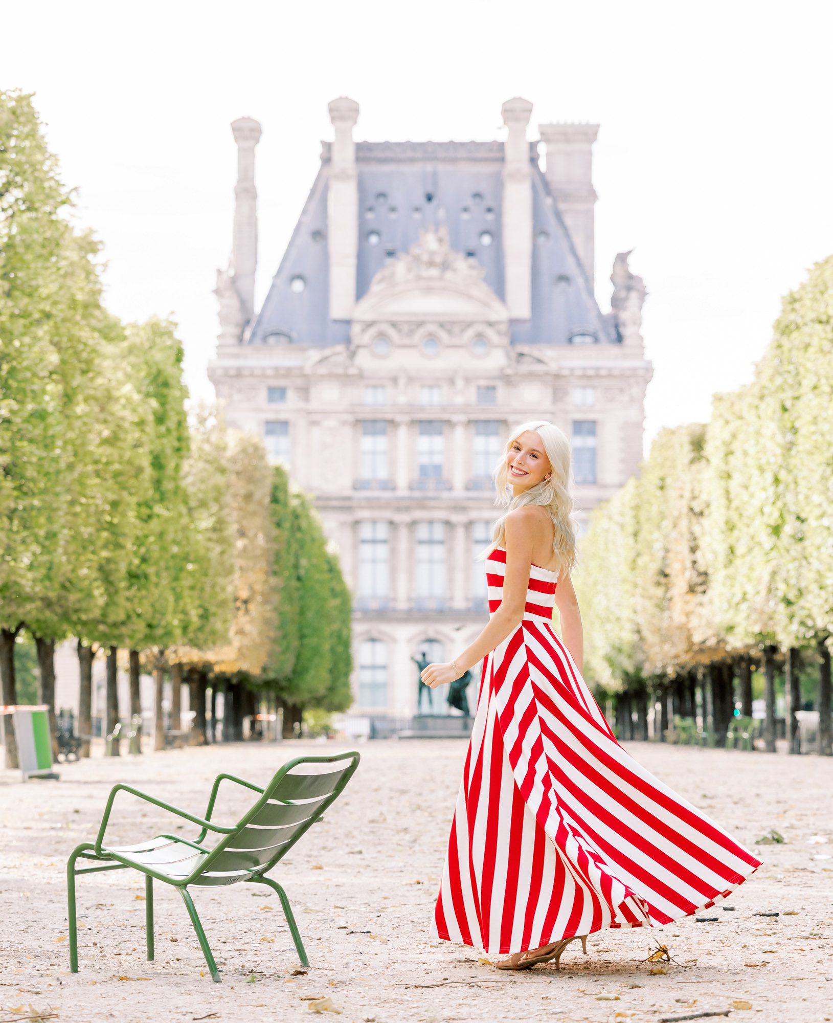 A high school senior girl in a red and white striped dress twirling during a graduation photoshoot in the Tuileries Garden. The Pavillon de Flore of the Louvre Museum and a classic green Parisian park chair are in the background.